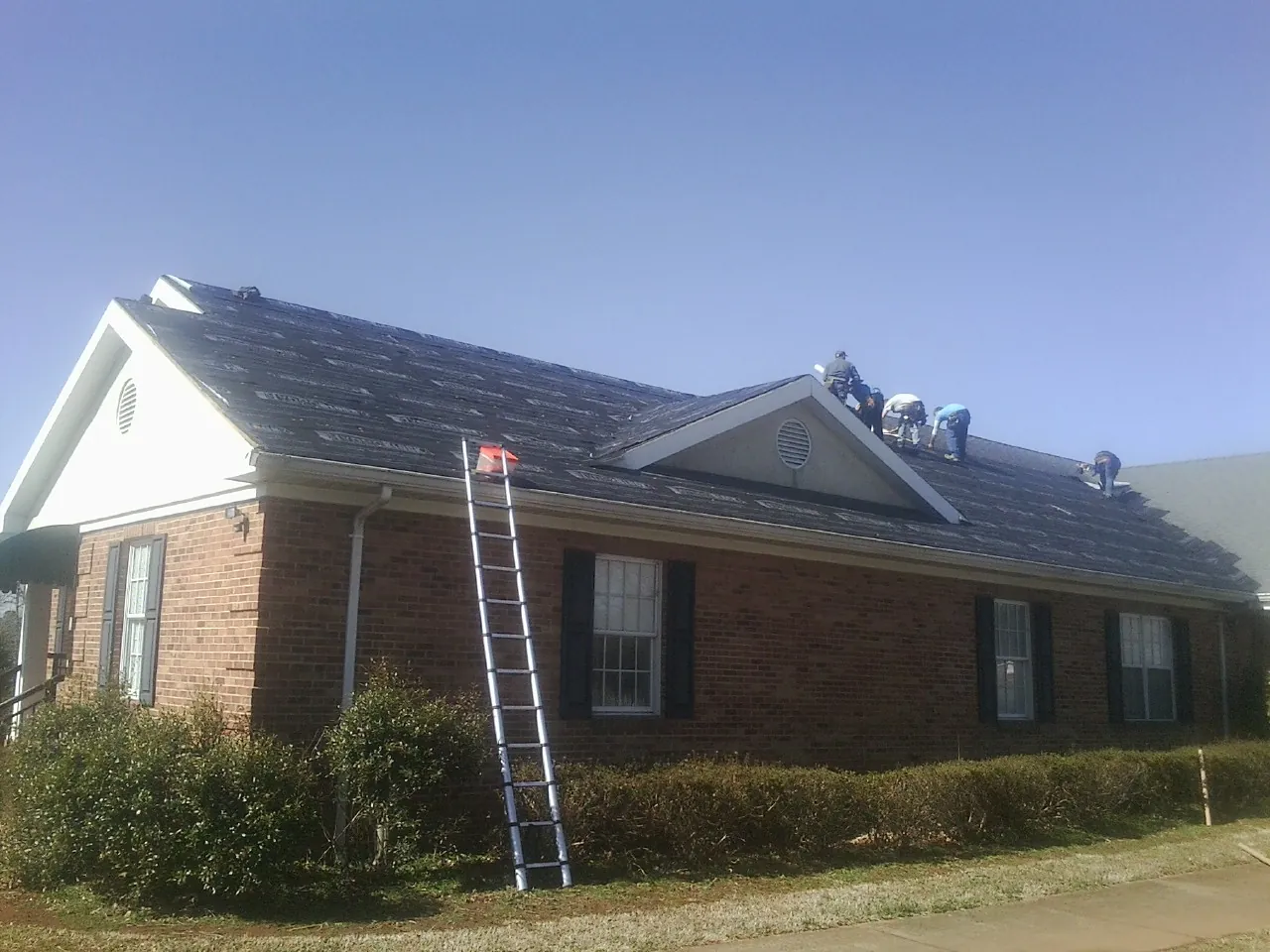 Crew tearing shingles off a brick ranch roof with a ladder set — Emergency Roof Repair work by our crew in East Hampstead