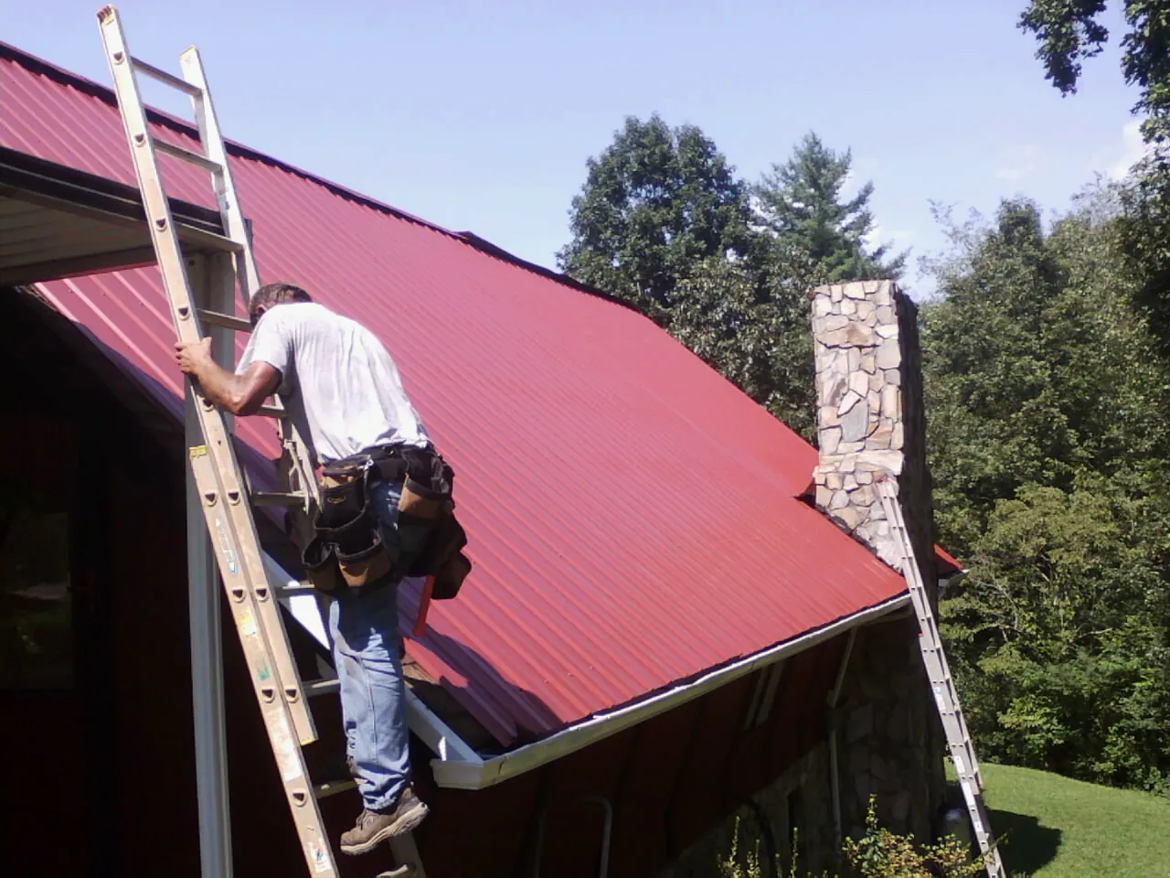 Roofer on a ladder beside a red metal roof and stone chimney — Soffit & Fascia Repair by Roofing Brothers in East Hampstead